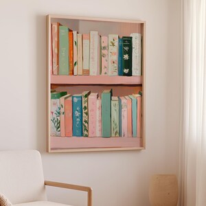 May include: A framed painting of a bookshelf filled with colorful books. The books are in shades of pink, green, blue, and orange, with floral and botanical designs. The bookshelf is painted pink and has a light wood frame.