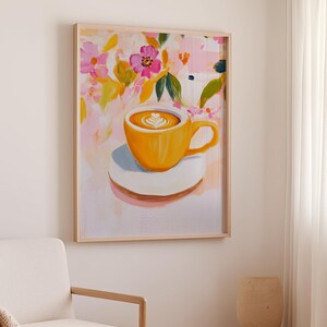 May include: A framed art print featuring a vibrant painting of a yellow coffee cup with latte art, resting on a white saucer. The background showcases a colorful floral design with pink, yellow, and green hues. The artwork is encased in a light-colored wooden frame.
