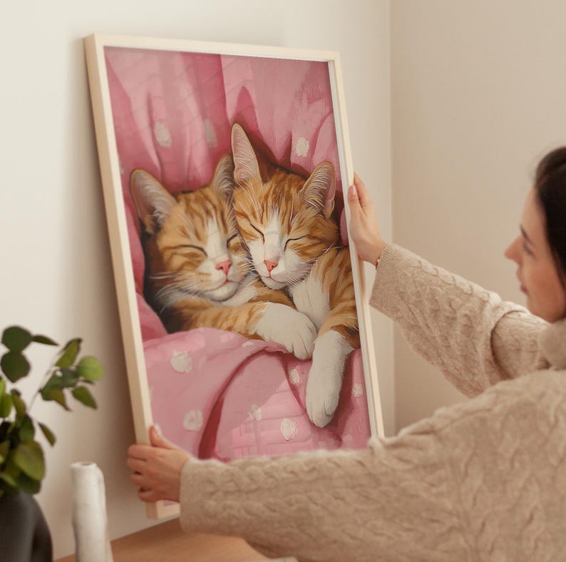 Op de afbeelding: Een ingelijst schilderij van twee oranje tabby-katten die knuffelen onder een roze deken met witte stippen.