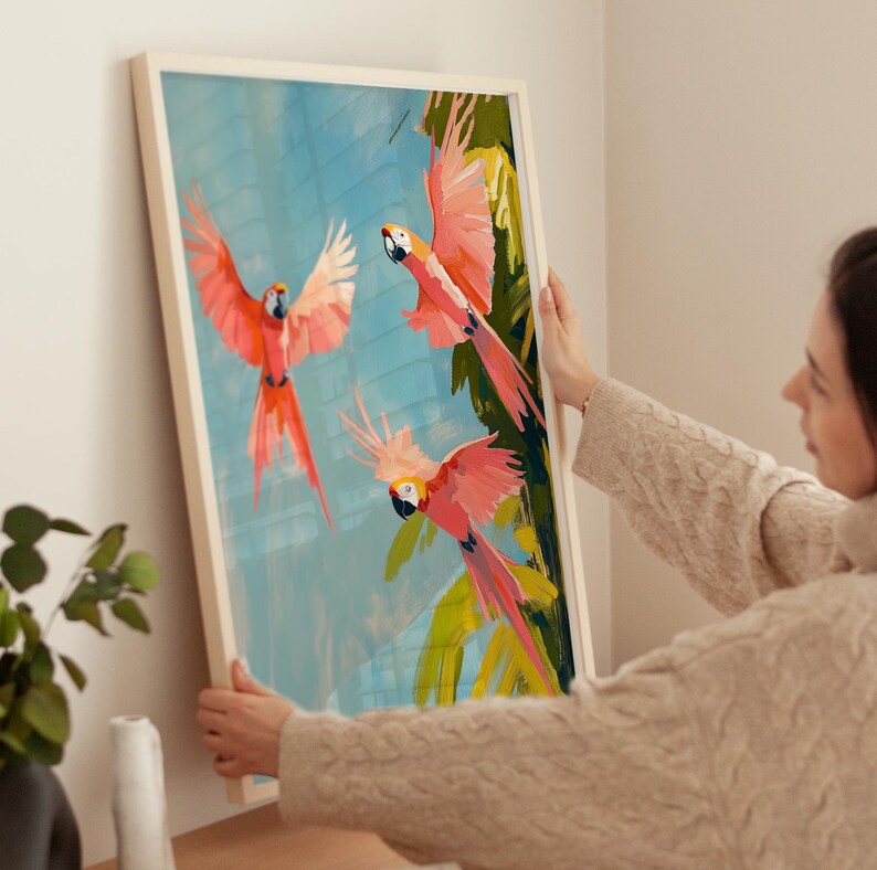 K&ouml;nnte beinhalten: Ein gerahmter Druck von drei rosa und roten Papageien, die in einer tropischen Umgebung mit gr&uuml;nen Bl&auml;ttern und blauem Himmel fliegen.
