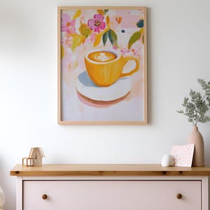 May include: A framed art print featuring a yellow coffee cup with latte art on a white saucer. The background has a floral design with pink, yellow, and green brushstrokes. The artwork is displayed on a light pink dresser.