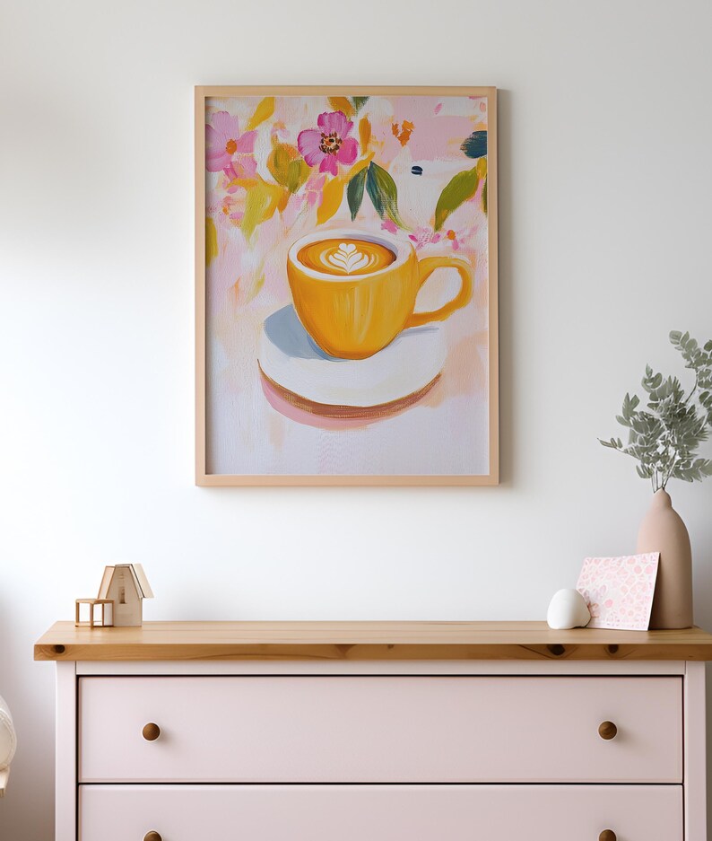 May include: A framed art print featuring a yellow coffee cup with latte art on a white saucer. The background has a floral design with pink, yellow, and green brushstrokes. The artwork is displayed on a light pink dresser.