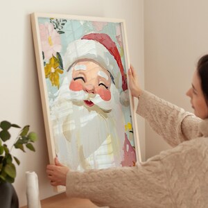May include: A framed painting of Santa Claus with a white beard and red hat. The painting has a whimsical style with a floral background in shades of pink, yellow, and blue.
