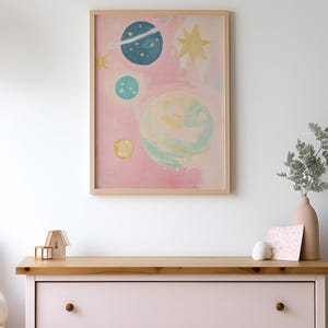 May include: A framed print of a pink watercolor painting with a whimsical depiction of planets and stars. The planets are painted in shades of blue, green, and yellow.