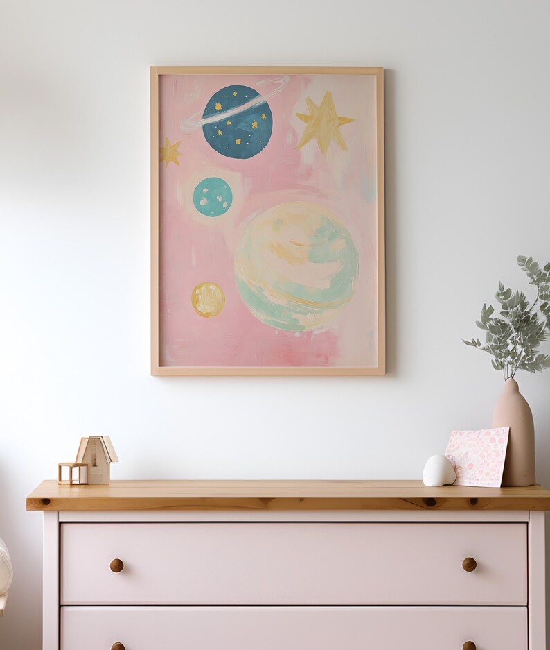 May include: A framed print of a pink watercolor painting with a whimsical depiction of planets and stars. The planets are painted in shades of blue, green, and yellow.