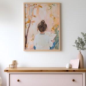 May include: A framed art print showing a person with brown hair in a bun, reading a book outdoors. The artwork uses a soft palette of pinks, yellows, and blues, with a light wood frame. The print is hanging above a pink chest of drawers.