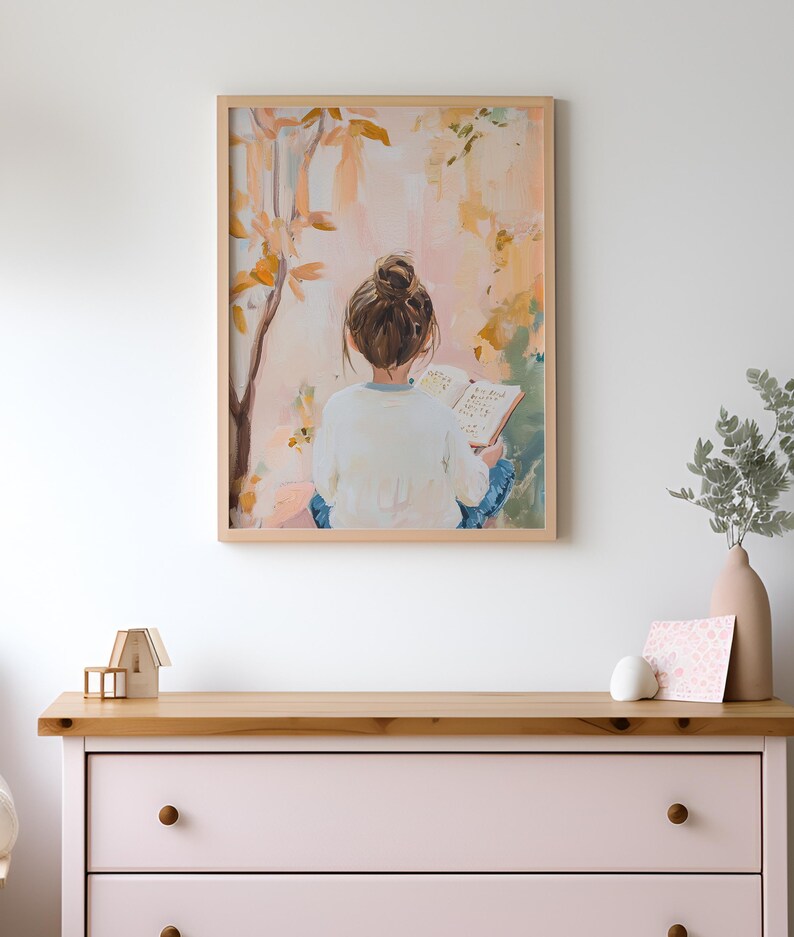 May include: A framed art print showing a person with brown hair in a bun, reading a book outdoors. The artwork uses a soft palette of pinks, yellows, and blues, with a light wood frame. The print is hanging above a pink chest of drawers.