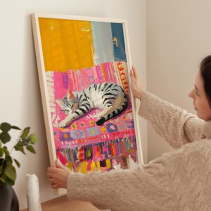 May include: A framed painting of a gray and white cat sleeping on a colorful rug. The rug has a pink, yellow, and blue pattern. The cat is curled up in a ball and has its eyes closed.