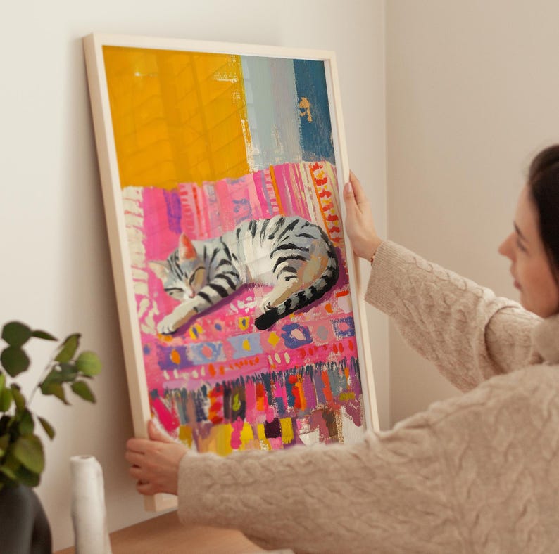 May include: A framed painting of a gray and white cat sleeping on a colorful rug. The rug has a pink, yellow, and blue pattern. The cat is curled up in a ball and has its eyes closed.