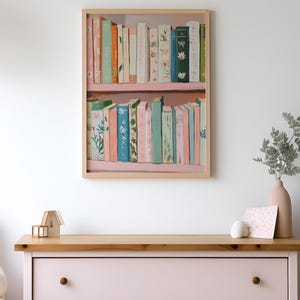 May include: A framed print of a bookshelf with colorful books. The books are in shades of pink, green, and orange, with floral and botanical designs. The frame is light wood, and the print is displayed above a pink dresser.