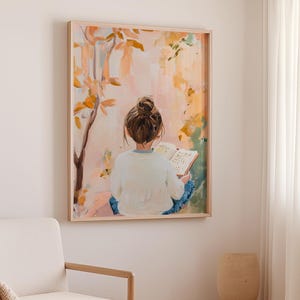 May include: A framed painting shows a person with a bun reading a book outside. The artwork uses a soft palette of pink, orange, and green, with a tree and foliage in the background. The frame is a light wood colour.