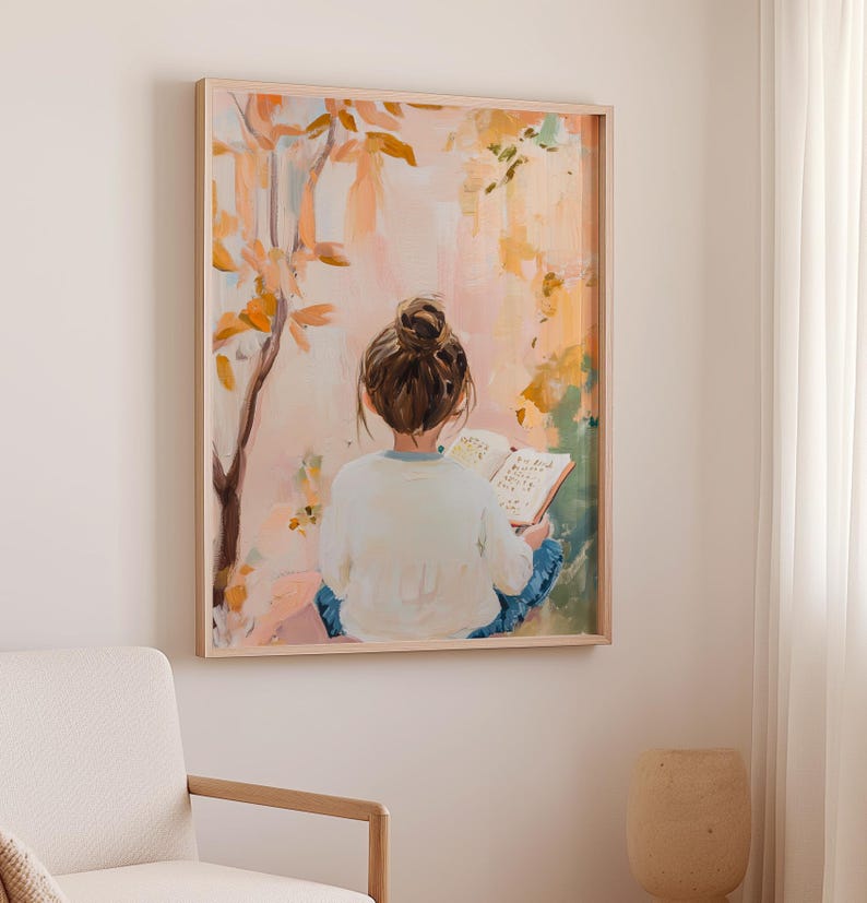 May include: A framed painting shows a person with a bun reading a book outside. The artwork uses a soft palette of pink, orange, and green, with a tree and foliage in the background. The frame is a light wood colour.