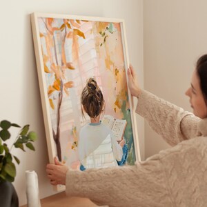 May include: A framed art print shows a person reading by a window, with a warm palette of orange, pink, and yellow. The artwork features a person with their back to the viewer, with a bun hairstyle, reading a book. The frame is a light beige colour.