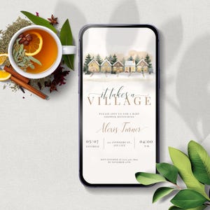 May include: A smartphone shows a baby shower invitation with the words "it takes a village." The invitation has a watercolour village illustration. A cup of tea with lemon and cinnamon is on the left, with other herbs.