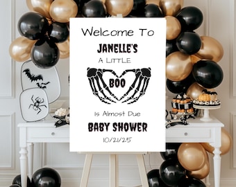 Pode incluir: Uma placa branca em um cavalete com o texto "Welcome To Janelle's A Little Boo Is Almost Due Baby Shower 10/21/25". A placa apresenta mãos de esqueleto formando um coração em torno da palavra "Boo". Balões pretos e dourados e decorações com tema de Halloween.