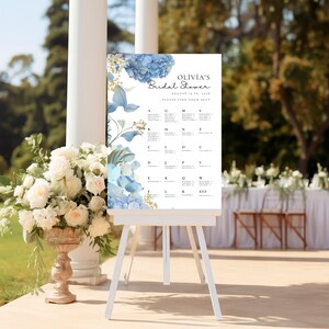 May include: A white sign with blue hydrangeas and gold accents on a white easel. The sign says "Olivia's Bridal Shower August 17th, 2019 Please Find Your Seat" and has a seating chart with letters of the alphabet.