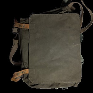 Puede incluir: Un bolso de mensajero de lona verde con correas y hebillas de cuero marrón. El bolso tiene un cierre de solapa y un pequeño bolsillo en la parte delantera. El bolso tiene la palabra "GRAFF" impresa en la correa.