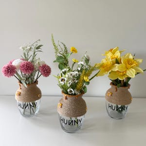 Può includere: Tre vasi di vetro con composizioni floreali. Ogni vaso è avvolto con spago e decorato con piccoli accenti di api gialle. La parola "HUNNY" è stampata in nero sulla parte anteriore di ogni vaso. I fiori includono fiori rosa, bianchi e gialli.