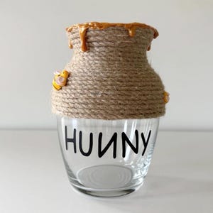 May include: A clear glass jar with the word HUNNY printed in black letters. The upper portion of the jar is wrapped in thick tan twine, with yellow and orange resin honey drips at the rim and a small yellow and black bee figurine attached.