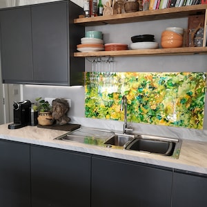 May include: A modern kitchen featuring dark grey cabinets, a stainless steel sink, and a marble worktop. The splashback is a colourful abstract design with green, yellow, and orange tones. There are shelves above the sink with various items, including books, dishes, and glasses.