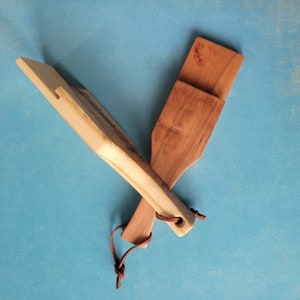 May include: Two wooden spatulas, one light brown and one dark brown, are crossed over each other. The light brown spatula has a leather strap attached to it.