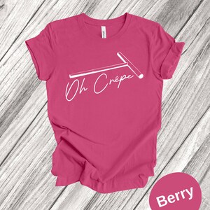 May include: A pink t-shirt with a white graphic of a crepe maker and the text "Oh Crepe". The shirt is on a wood background. The text "Berry" is in a circle below the shirt.