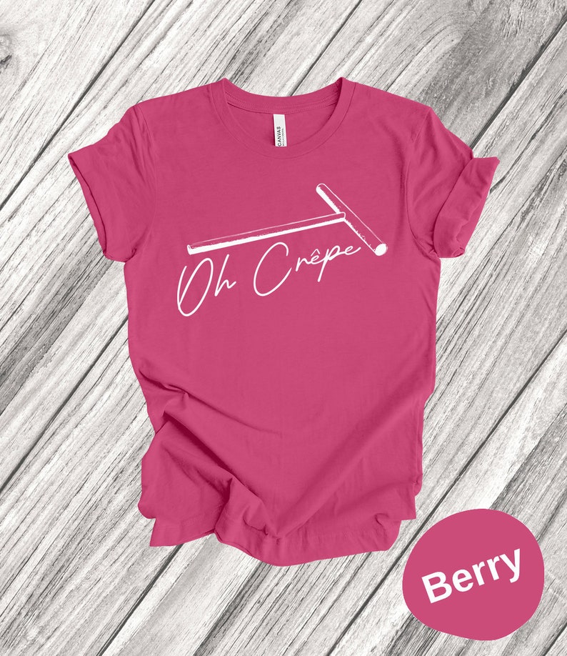 May include: A pink t-shirt with a white graphic of a crepe maker and the text "Oh Crepe". The shirt is on a wood background. The text "Berry" is in a circle below the shirt.