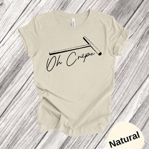 May include: A beige t-shirt with a black graphic of a crepe maker and the text "Oh Cr&ecirc;pe". The shirt is on a wooden background. The colour of the shirt is "Natural".