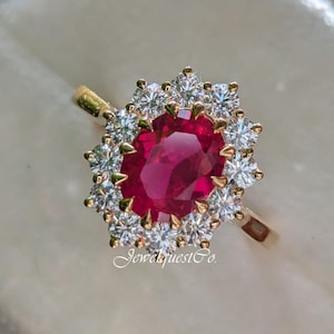 Può includere: Un anello d'oro con un grande rubino ovale circondato da diamanti più piccoli. L'anello è in una scatola per anelli bianca.