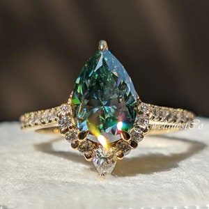 May include: A pear-shaped green moissanite engagement ring with a halo of smaller diamonds set in a yellow gold band. The ring is displayed in a white ring box.