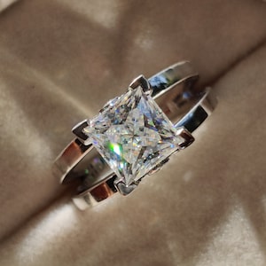 Könnte beinhalten: Ein silberner Ring mit einem quadratischen Diamanten in der Mitte. Der Ring befindet sich in einer weißen Ringbox.