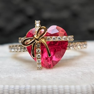 Könnte beinhalten: Ein goldener Ring mit einem herzförmigen roten Edelstein und einer goldenen Schleife oben. Der Ring ist mit kleinen klaren Edelsteinen um das Herz herum besetzt.