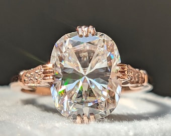 Puede incluir: Anillo de compromiso de oro rosa con un gran diamante de talla ovalada. El anillo tiene una banda delicada con detalles intrincados en los lados. El diamante brilla intensamente sobre un fondo gris suave. Una joya clásica y elegante.