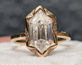 Puede incluir: Un anillo de oro con una gran piedra preciosa transparente de corte hexagonal alargado como piedra central. El engaste está hecho de oro amarillo pulido e incluye pequeñas piedras de acento en pavé a lo largo de los bordes puntiagudos.