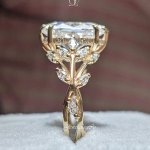 May include: A gold ring with a large, clear, rectangular-cut gemstone. The ring features a detailed floral design with small accent stones. The band is intricately designed, and the ring is set on a light blue surface.