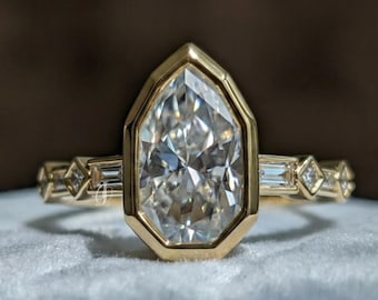 Puede incluir: Un anillo de oro con una gran piedra central en forma de pera en un engaste de bisel. La banda está decorada con piedras alternas de corte baguette y corte cuadrado pequeño a cada lado, creando un diseño simétrico con acabado pulido.