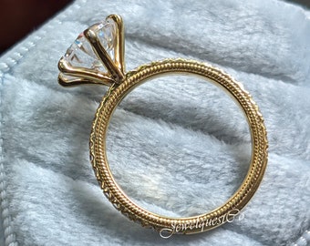 May include: A gold engagement ring with a large, round, clear diamond set in a prong setting. The ring band is decorated with a delicate, braided design.