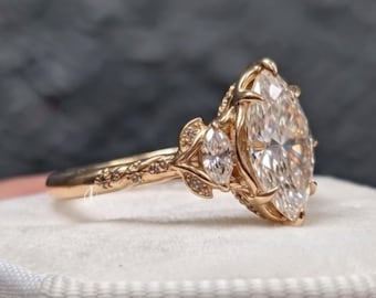 Puede incluir: Un anillo de oro rosa con una gran piedra central de talla marquesa flanqueada por piedras laterales más pequeñas de talla marquesa. La banda presenta un intrincado trabajo de metal en forma de hojas y pequeñas piedras engastadas en pavé.