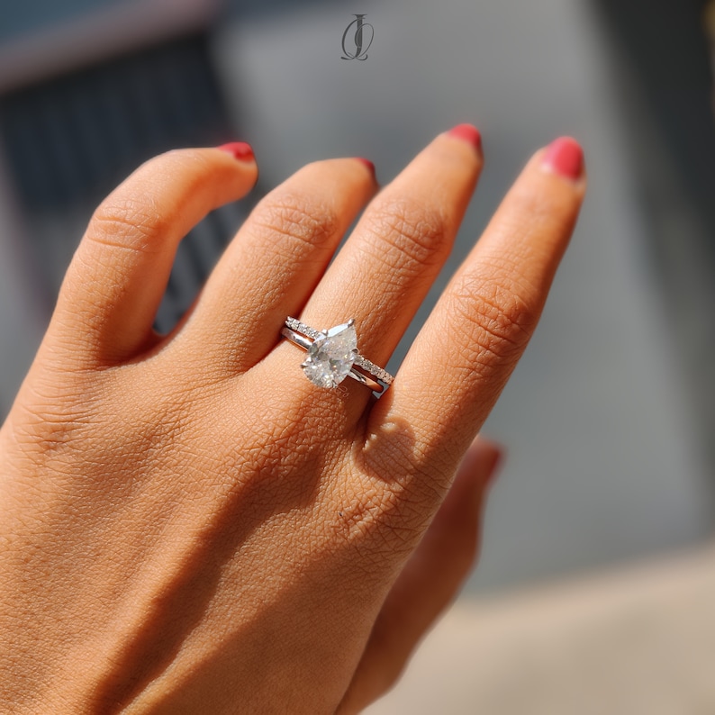 May include: A pear-shaped diamond engagement ring and a matching band are displayed on a hand with red nail polish. The rings are set in a silver metal, and the main ring features a large, clear pear-cut diamond.
