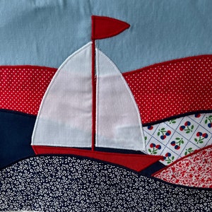 May include: A white sailboat with a red sail and a red hull is appliqued on a blue fabric background. The background is made of different patterned fabrics, including red with white polka dots, white with red and blue cherries, and white with red flowers.