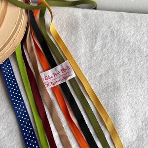 Op de afbeelding: Assortiment van lintrollen in verschillende kleuren, waaronder groen, geel, oranje, rood, zwart en blauw. De linten zijn waaiervormig gerangschikt en hebben een wit label met de tekst "The Red Mill Collection by Karol Lynne".