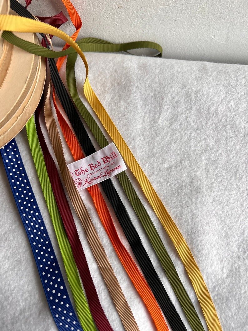 Op de afbeelding: Assortiment van lintrollen in verschillende kleuren, waaronder groen, geel, oranje, rood, zwart en blauw. De linten zijn waaiervormig gerangschikt en hebben een wit label met de tekst "The Red Mill Collection by Karol Lynne".