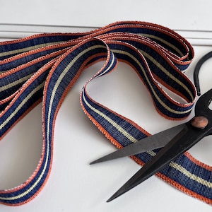 May include: A close-up of a spool of navy blue and cream striped ribbon with orange edges. A pair of black fabric shears are lying on top of the ribbon.