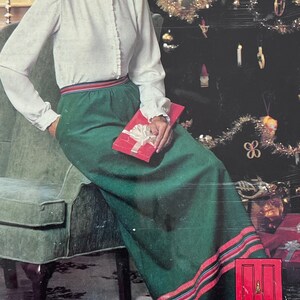 Op de afbeelding: Een vrouw in een wit blouse en een groene rok met rode en blauwe strepen zit op een stoel voor een gedecoreerde kerstboom. De afbeelding is afkomstig uit een kerstcatalogus van 1979 voor The Talbot, een kledingwinkel.