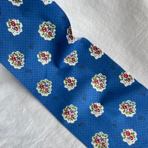 May include: A blue fabric with a repeating floral pattern. The flowers are red, purple, and white, with green leaves. The background is a solid blue with small black dots. The fabric is folded over a white surface.