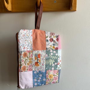 May include: A patchwork fabric bag with a brown leather handle and zipper. The bag features a variety of floral and polka dot patterns in shades of pink, orange, blue, and green. The word "yoyo" is printed on a small label.