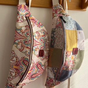 May include: Two fabric fanny packs hanging on a wooden rack. One is a paisley print in pink, blue, and yellow. The other is a patchwork design in pink, blue, yellow, and green. Both have a brown zipper and a white strap. The text "Musmutlu günlerimiz olsunn" is written above the bags.