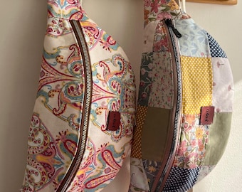 May include: Two fabric fanny packs hanging on a wooden rack. One is a paisley print in pink, blue, and yellow. The other is a patchwork design in pink, blue, yellow, and green. Both have a brown zipper and a white strap. The text "Musmutlu günlerimiz olsunn" is written above the bags.