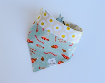 May include: A turquoise blue dog bandana with a print of bacon, eggs, and other breakfast foods. The bandana has a cream-colored trim with a print of yellow eggs with smiley faces. The bandana has a small label that says "Mutty Made" on it.