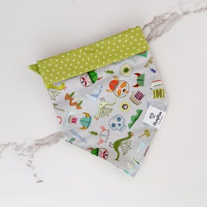 May include: A green and white bandana with a pattern of dragons, skulls, and viking helmets. The bandana has a green border with white polka dots and the text "Matty & Milo" in a white circle.